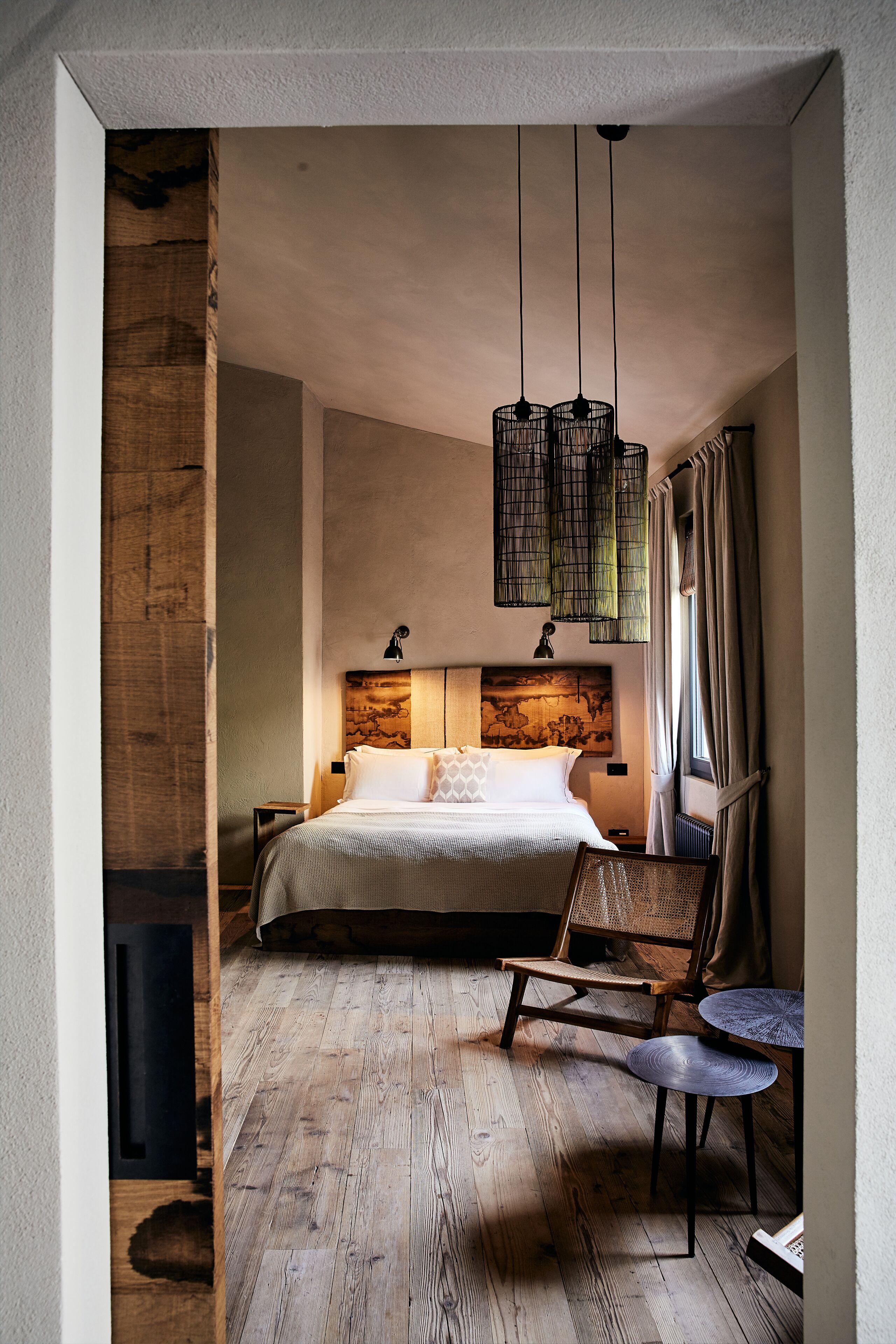 Un dormitorio con piso de madera, una cama con cabecera, una silla, una mesita y lámparas colgantes.