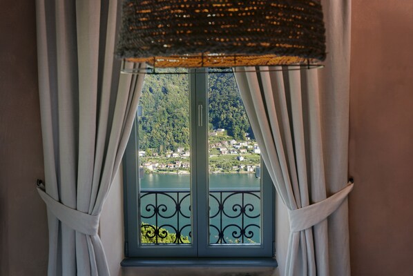 A window with white curtains and a metal railing, offering a view of a lake and a hillside with buildings. - Relais Castello di Morcote (Vico Morcote)