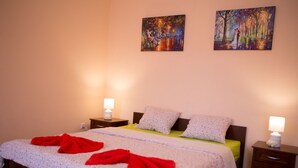 Een netjes opgemaakt bed met een bloemenbedekking en rode handdoeken, twee bedlampen en twee schilderijen aan de muur.