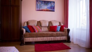 Een woonkamer met een bruine bank, rode en kleurrijke kussens, een rood tapijt, een houten kast en een schilderij aan de muur.
