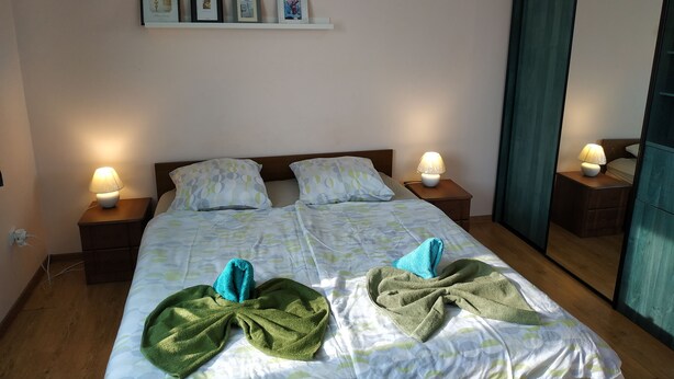 A neatly made bed with a patterned bedspread, two bedside lamps, and a wall with framed pictures and a small shelf.