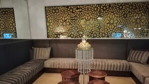 Un salon moderne avec un canapé d’angle, une table basse centrale en verre et quatre tabourets, ainsi qu’un panneau décoratif mural.