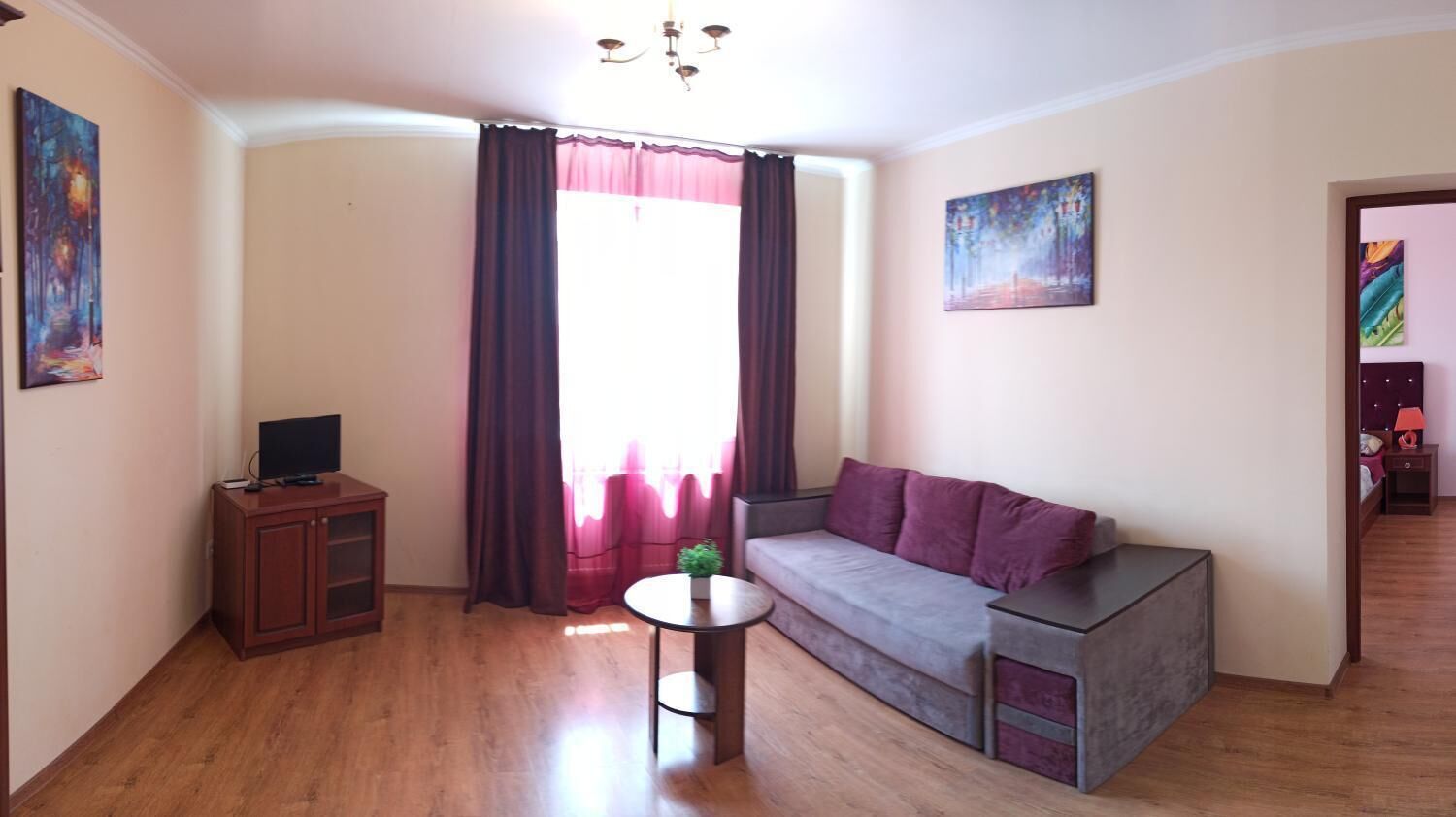 A living room with a sofa, a small table, a TV on a cabinet, and a window with curtains.