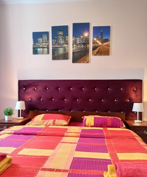 Une chambre d’hôtel avec un grand lit, une tête de lit sombre et trois tableaux représentant des paysages urbains accrochés au mur.