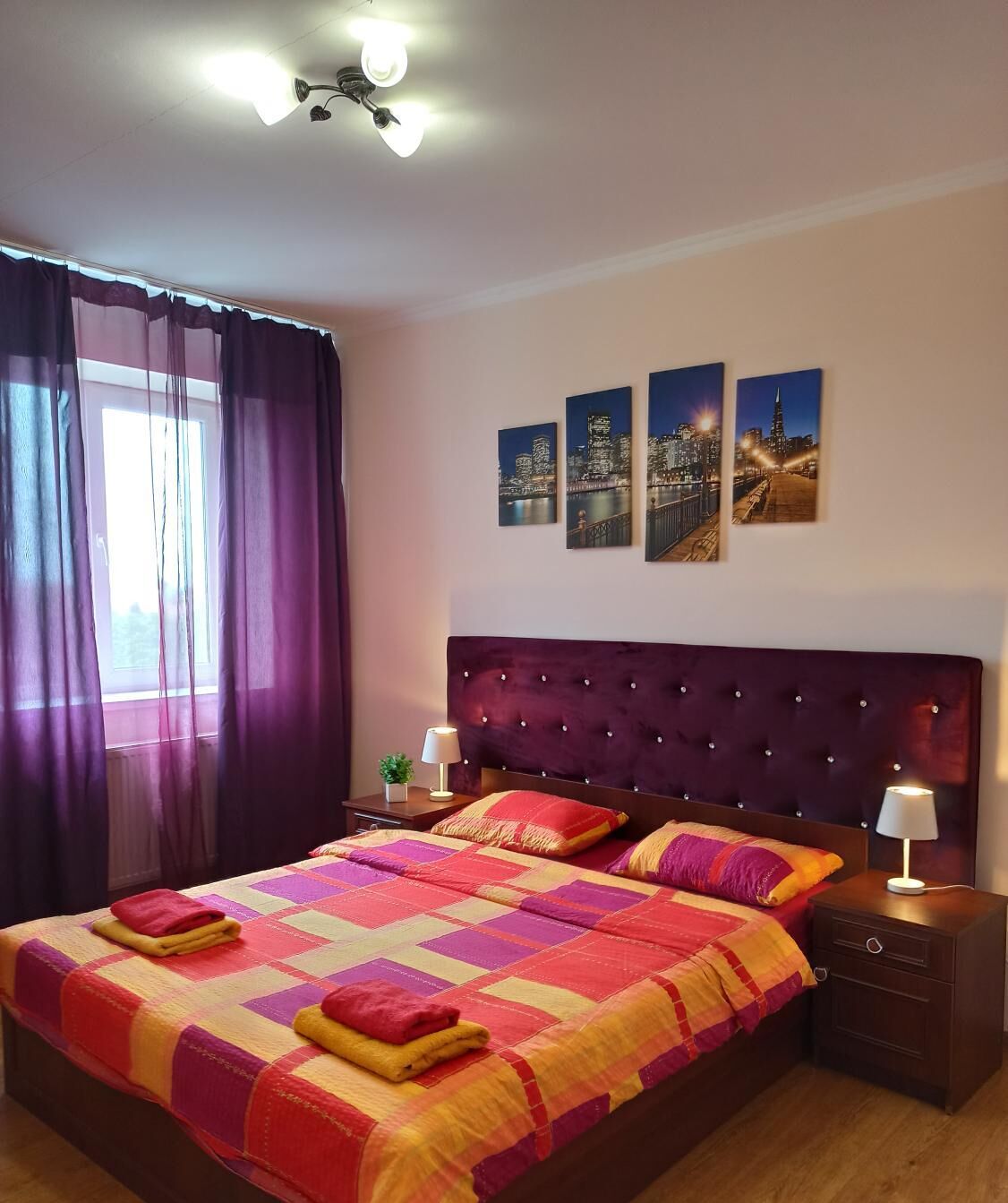 A hotel room with a large bed, a headboard with buttons, a nightstand with a lamp, and a window with purple curtains.