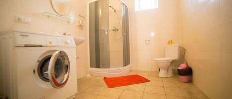 A bathroom with a washing machine, a glass shower enclosure, a toilet, and a red bath mat.