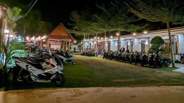 A nighttime view of a hotel with rows of parked motorcycles, a covered pavilion, and a building with multiple windows. - Rivana Hotel (Cijulang)
