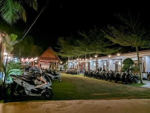 A nighttime view of a hotel with rows of parked motorcycles, a covered pavilion, and a building with multiple windows. - Rivana Hotel (Cijulang)