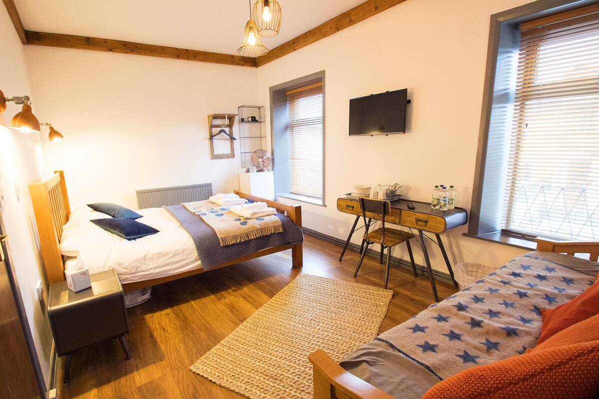 A bedroom with a bed, a desk, a chair, a TV, and a window with blinds.