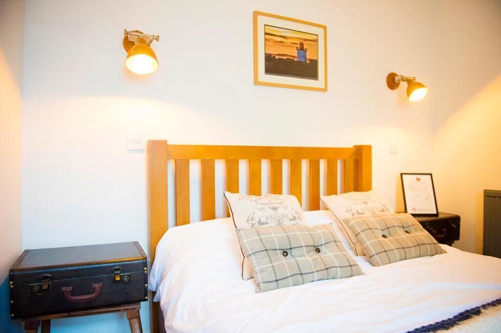 A bedroom with a wooden headboard, a bed with white bedding, two bedside tables, a suitcase, and wall-mounted lamps.