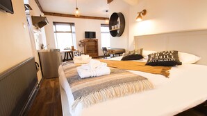 A hotel room with a large bed, a wooden floor, a television, a fan, and a shelf with decorative items. - Nik The Greek (Burry Port)
