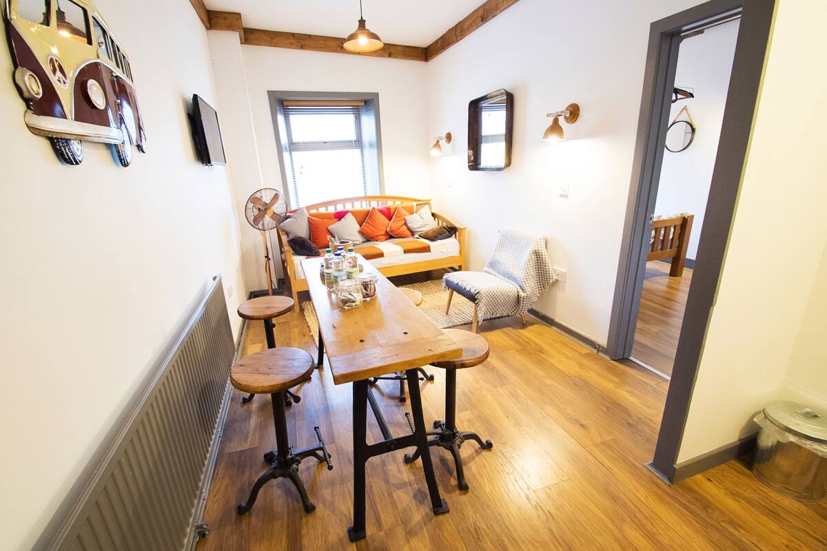 A cozy room with a wooden dining table, a sofa with colorful cushions, and a large window.