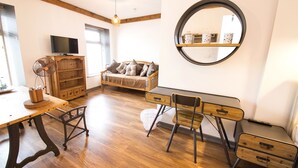 A room with a wooden floor, a round mirror, a TV, a sofa, a desk with drawers, and a fan. - Nik The Greek (Burry Port)