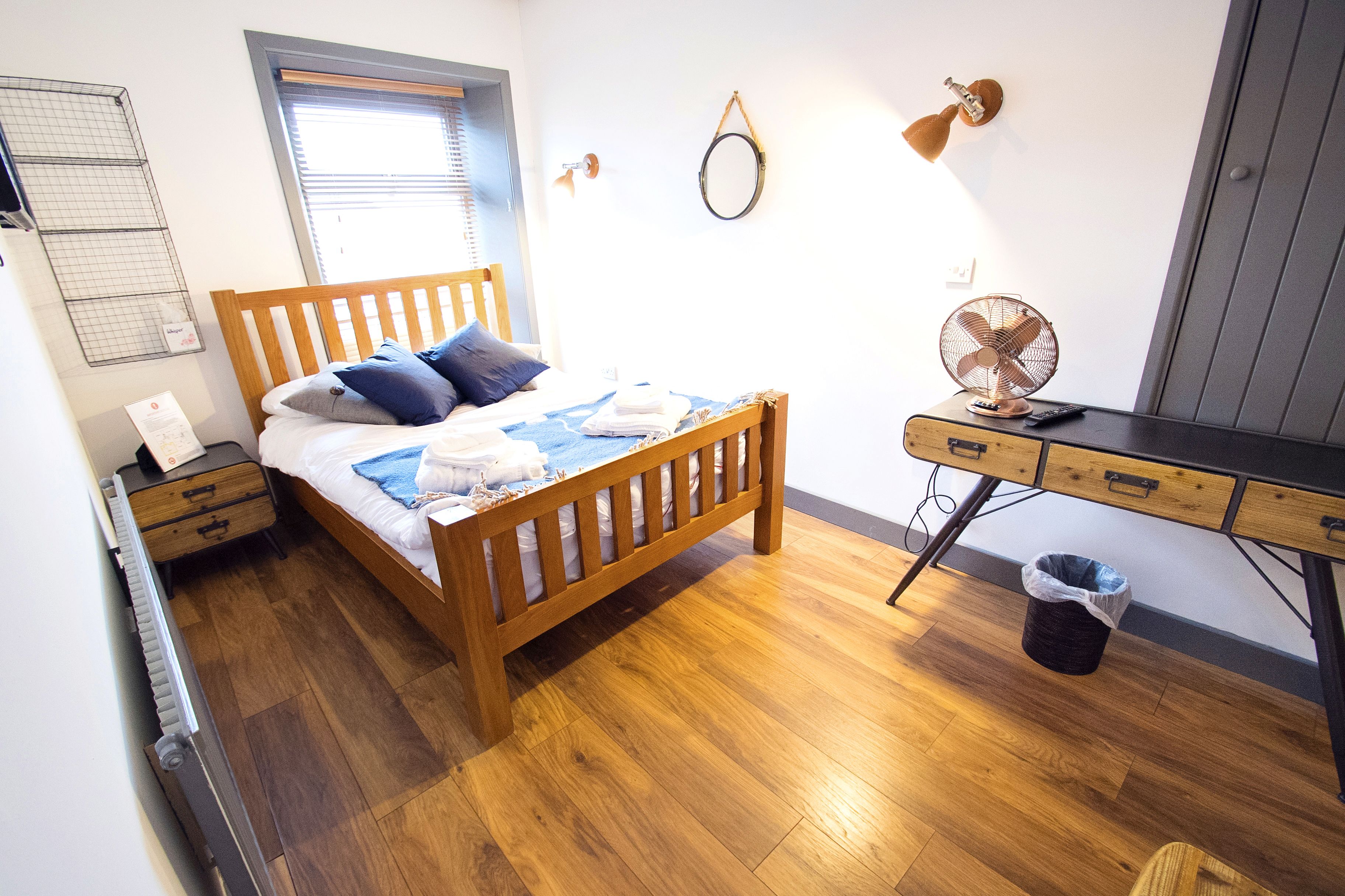 A bedroom with a wooden bed, a desk, a wall-mounted fan, a waste bin, and a window with blinds.
