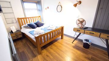 A bedroom with a wooden bed, a desk, a wall-mounted fan, a waste bin, and a window with blinds.