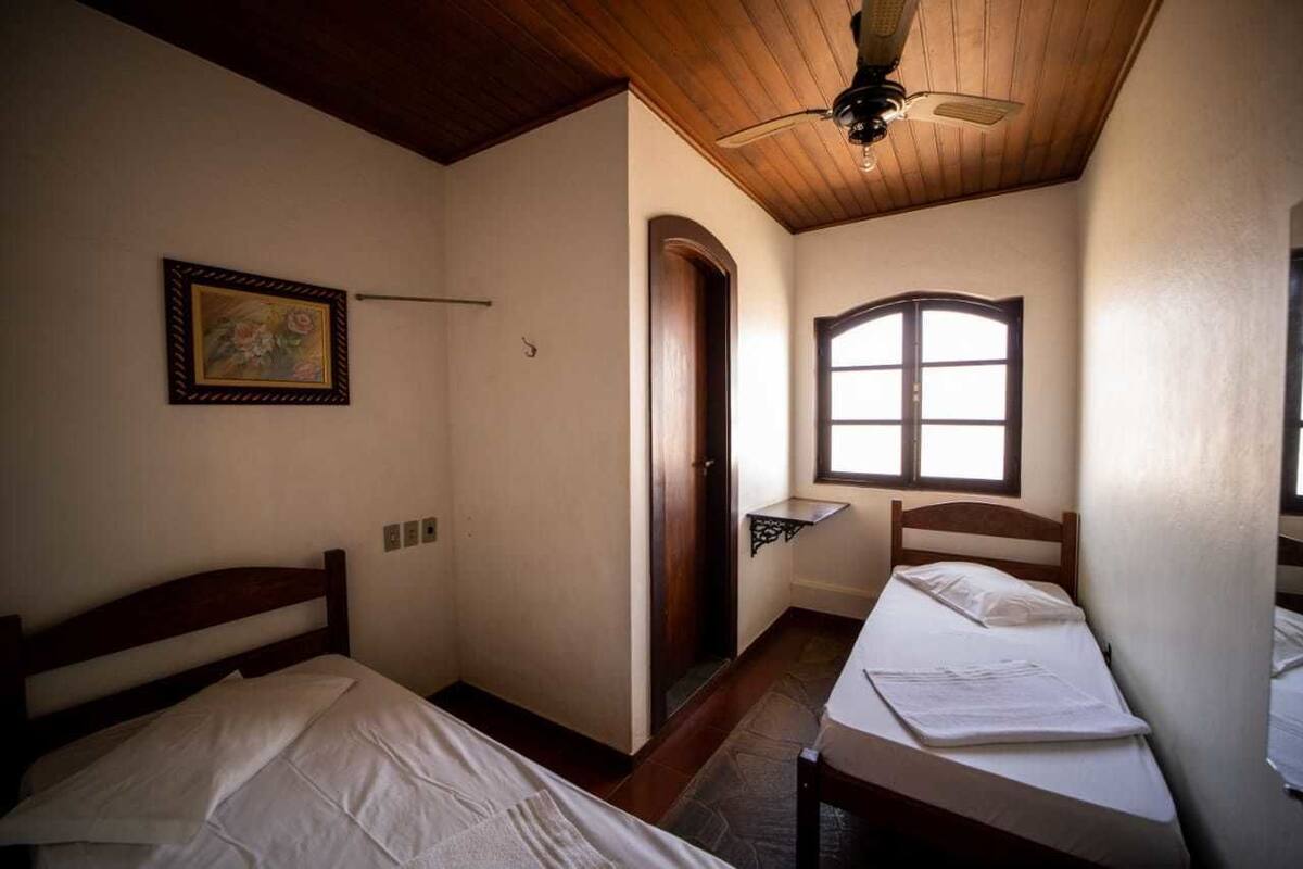 Une petite chambre accueillante, dotée de deux lits simples, d’un ventilateur de plafond en bois, d’un tableau encadré au mur et d’une fenêtre laissant entrer la lumière naturelle.