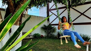 Une personne est assise sur une balançoire dans un jardin, avec un mur blanc et des poutres en bois en arrière-plan.