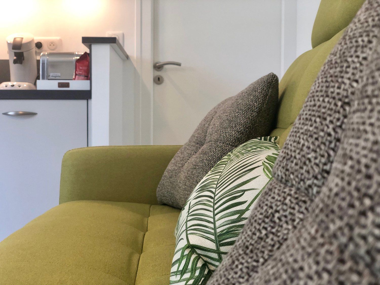 A green sofa with multiple cushions, a coffee machine, and a small cabinet in the background.