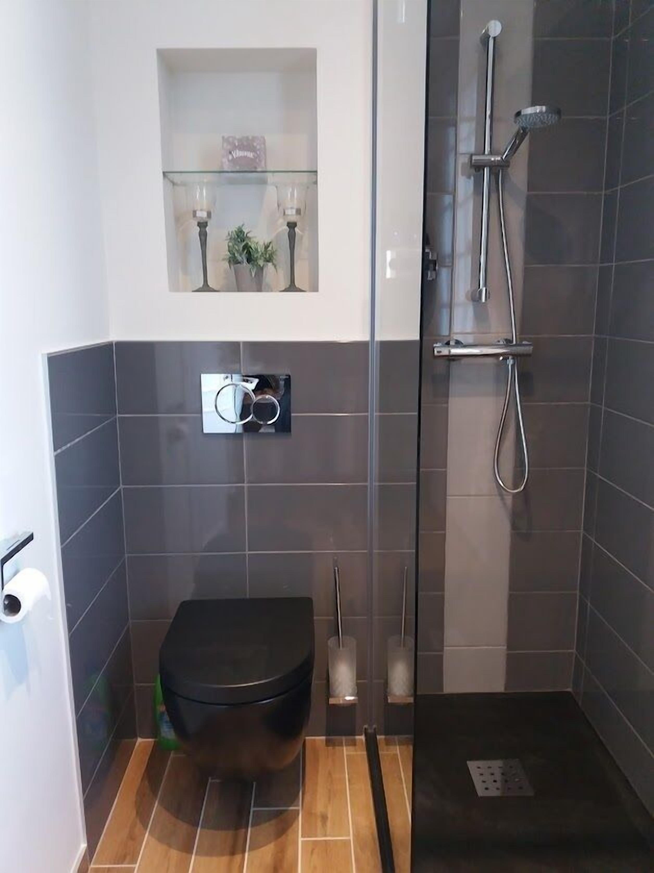 A compact bathroom with a toilet, a glass shower enclosure, and a small shelf with decorative items.