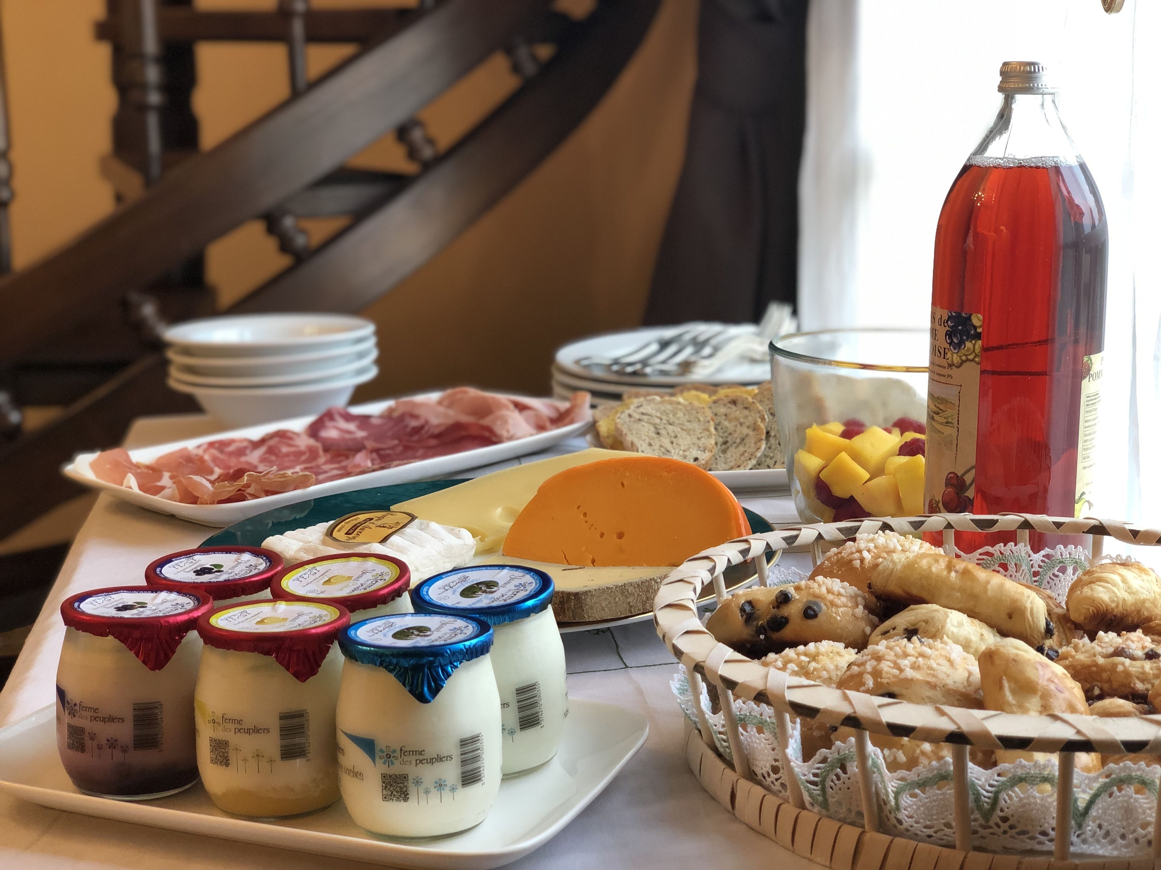 A breakfast buffet with various cheeses, meats, pastries, and a bottle of red wine.