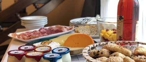 A breakfast buffet with various cheeses, meats, pastries, and a bottle of red wine.