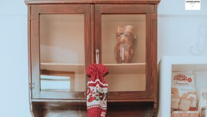 Ein Holzkabinett mit Glastüren, ein Stoff mit roten und weißen Mustern, der daran hängt, und eine Keramik-Eule. Darunter befinden sich Regale mit Büchern, eine kleine Holzschatulle und ein gerahmtes Bild.