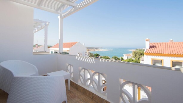 A balcony with a white wicker chair and table, overlooking a coastal view with buildings and a clear sky.
