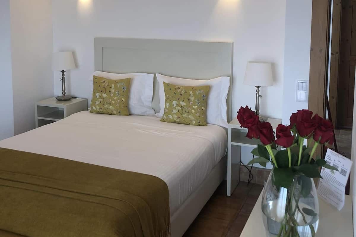A bedroom with a large bed, two bedside tables with lamps, a wooden ceiling, and a vase with flowers on a nightstand.