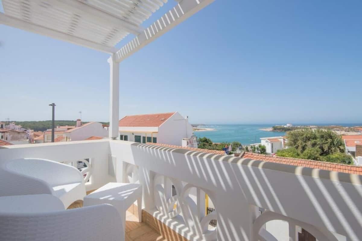 A balcony with white furniture and a view of the sea and buildings.