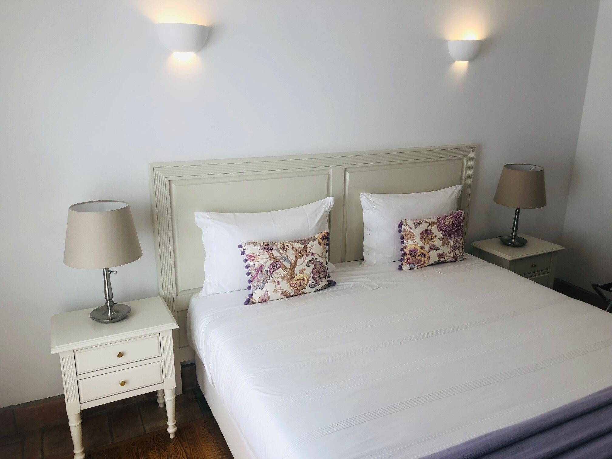 Een net opgemaakt bed met witte lakens en kussens met bloemenprint, aan weerszijden geflankeerd door nachtkastjes met lampen, in een eenvoudige kamer met lichtgekleurde muren en een houten vloer.