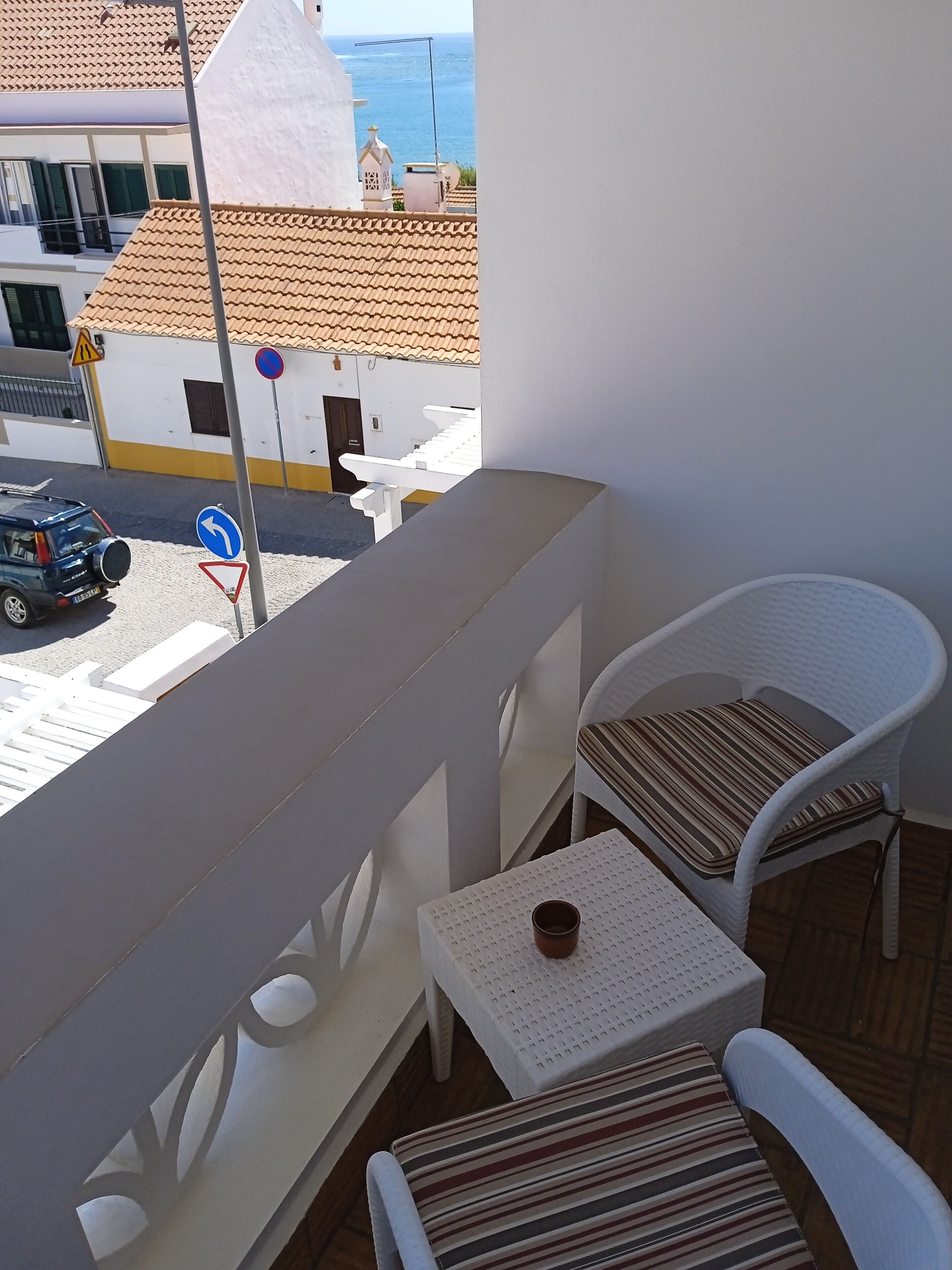 A balcony with white chairs and a small table, overlooking a street with a blue car and a building with a tiled roof.