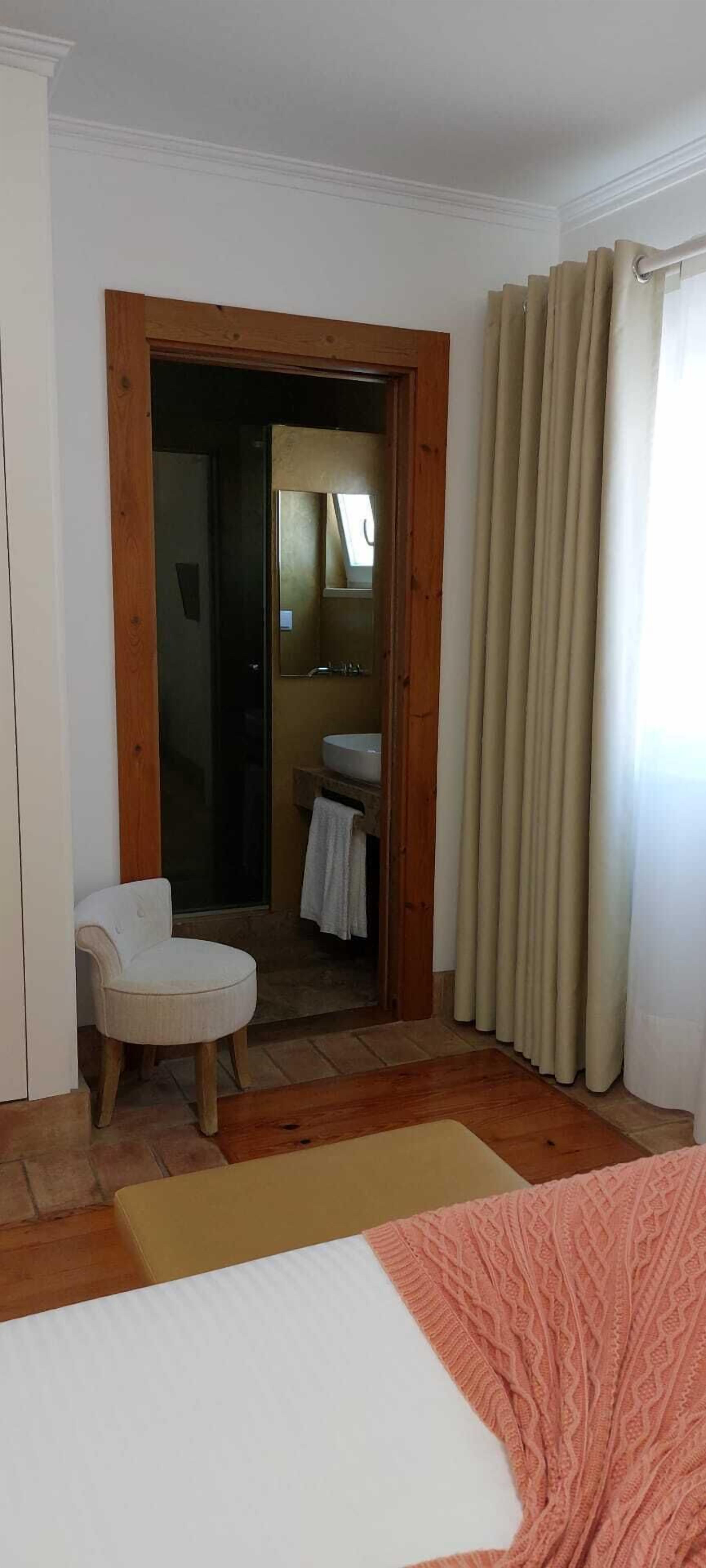 Une chambre à coucher avec un lit, une chaise, une porte en bois donnant sur une salle de bain et des rideaux de couleur beige.