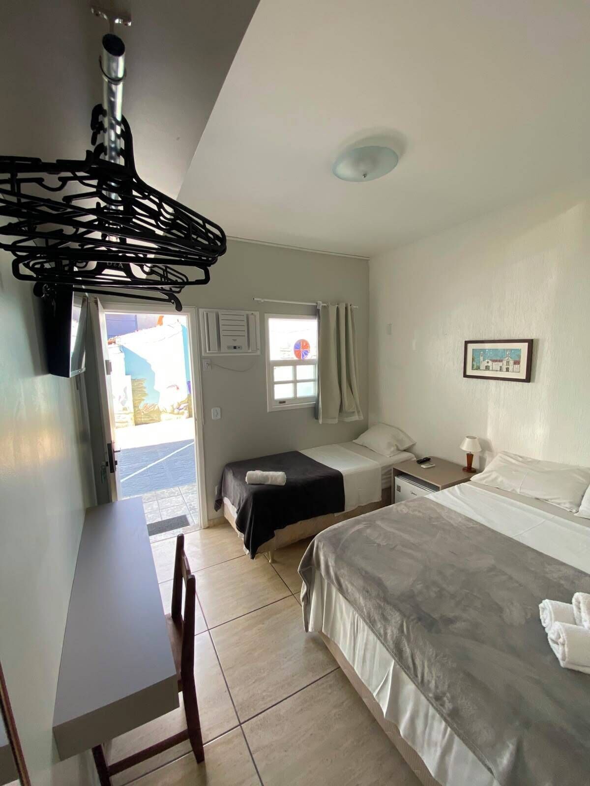 Quarto de hotel com cama, escrivaninha, cadeira, janela com cortinas, ar condicionado fixado na parede e pequena mesa com abajur.