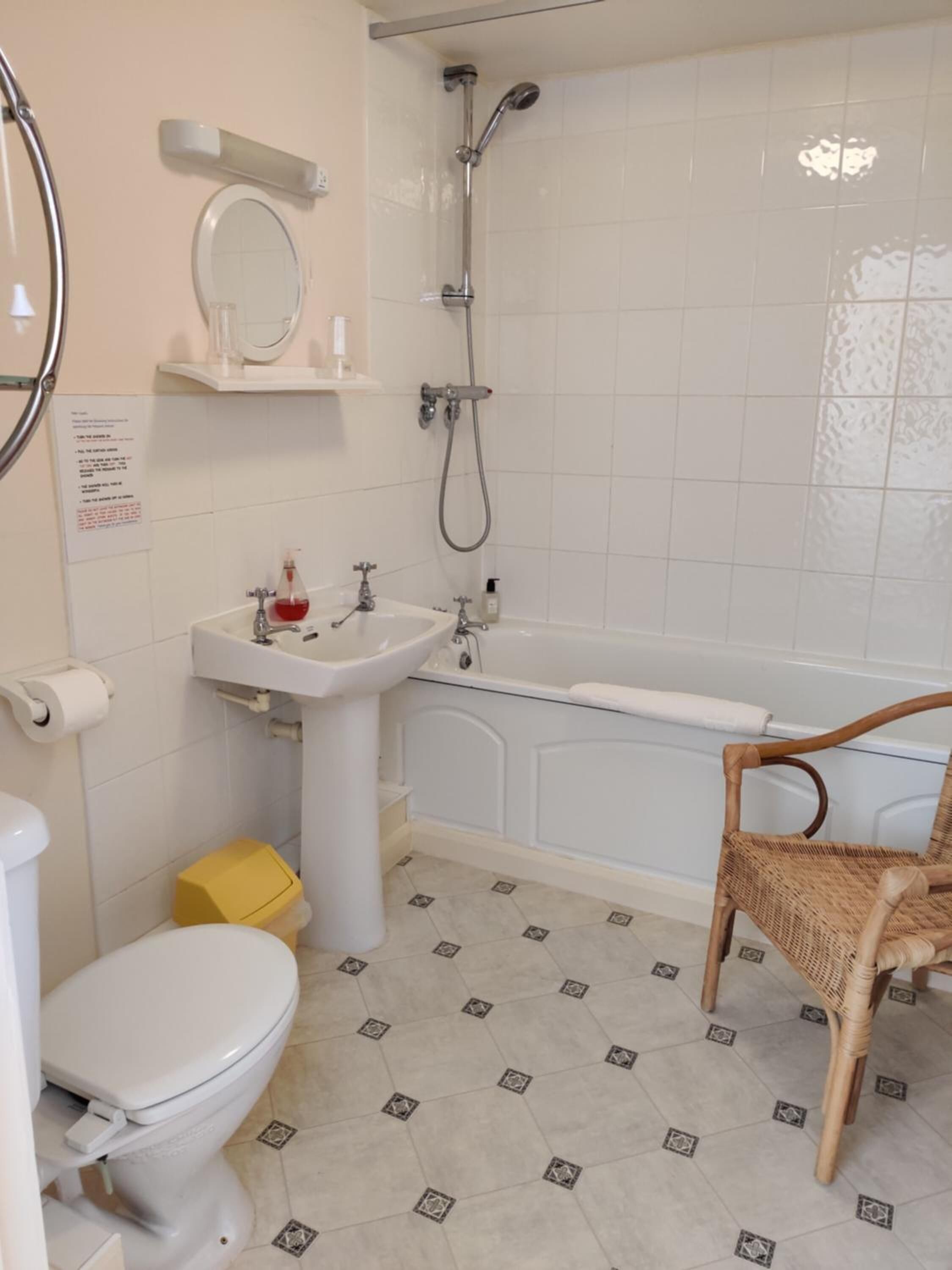 A bathroom with a toilet, sink, bathtub, shower, and a wicker chair.