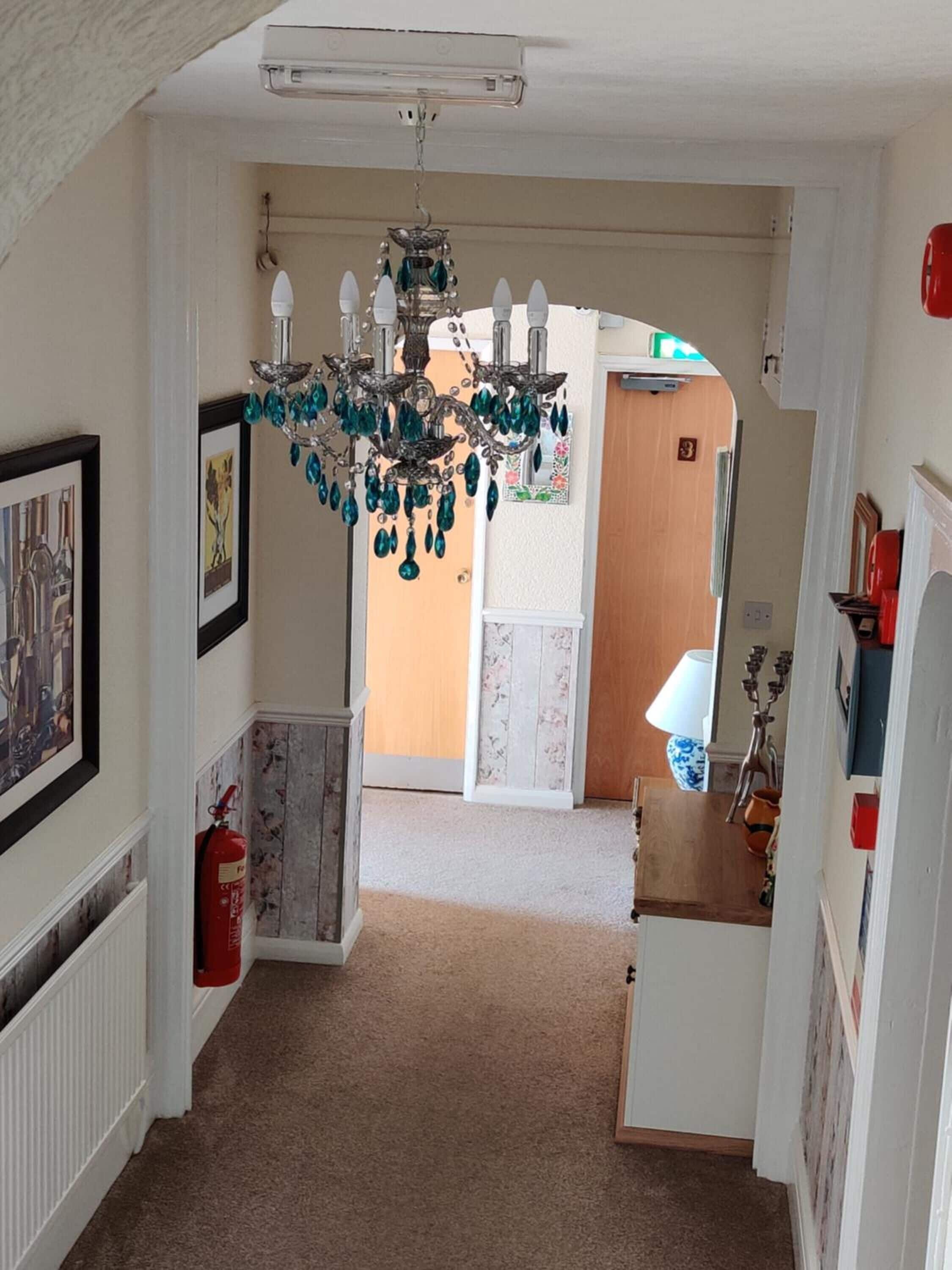 A hallway with a chandelier, framed pictures, a fire extinguisher, and a door leading to another room.