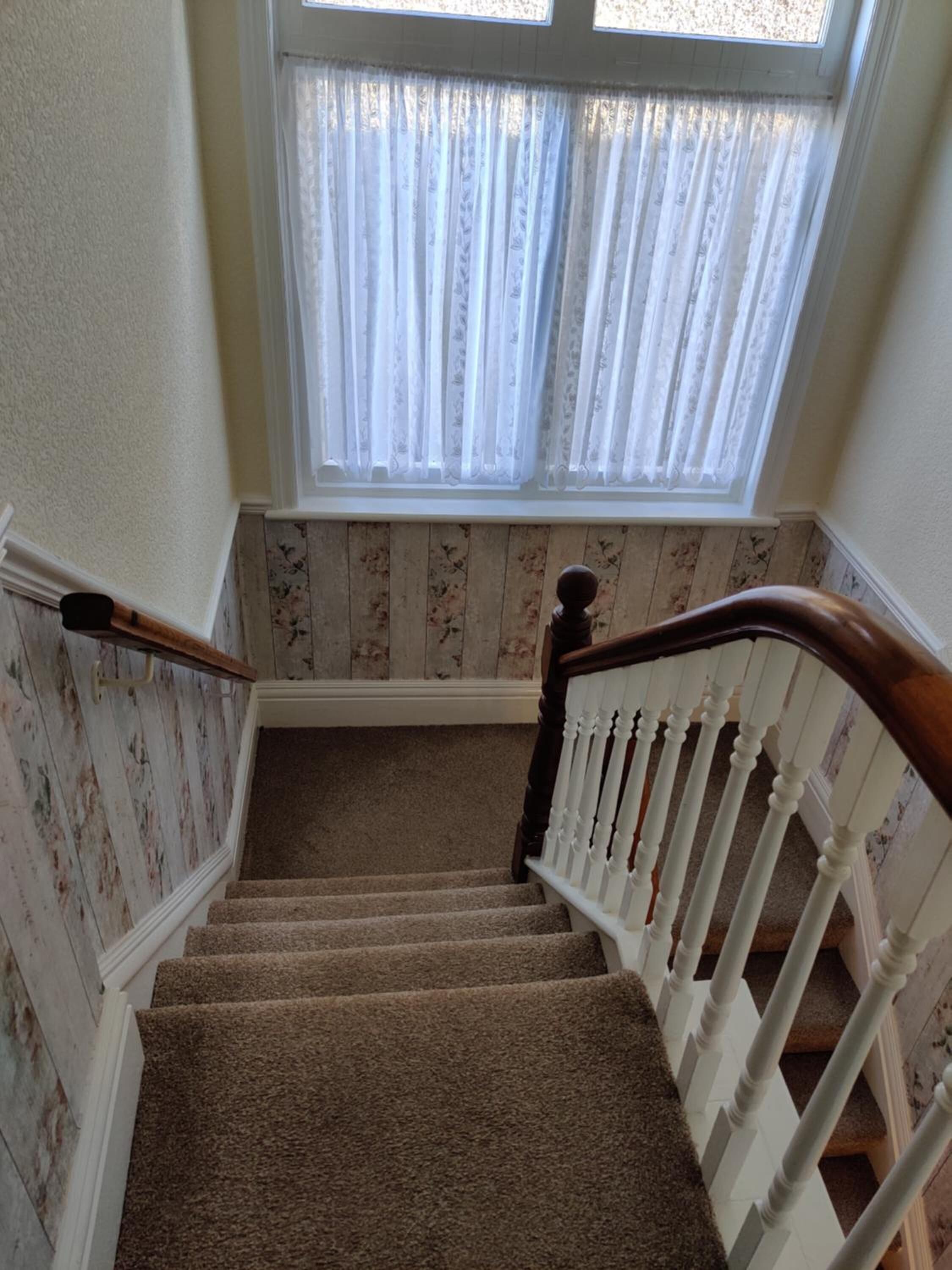 Un escalier recouvert de moquette, avec des balustrades blanches et une main courante en bois, menant à une pièce dont les murs sont recouverts d’un papier peint à motifs floraux et qui possède une fenêtre ornée de rideaux blancs.