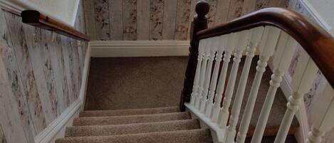 A carpeted staircase with white balusters and a wooden handrail, leading to a room with floral wallpaper and a window with white curtains.