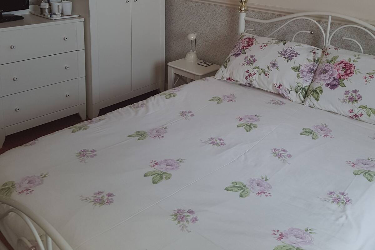 A bedroom with a bed, a wardrobe, a nightstand, and a desk with a computer and a coffee maker.