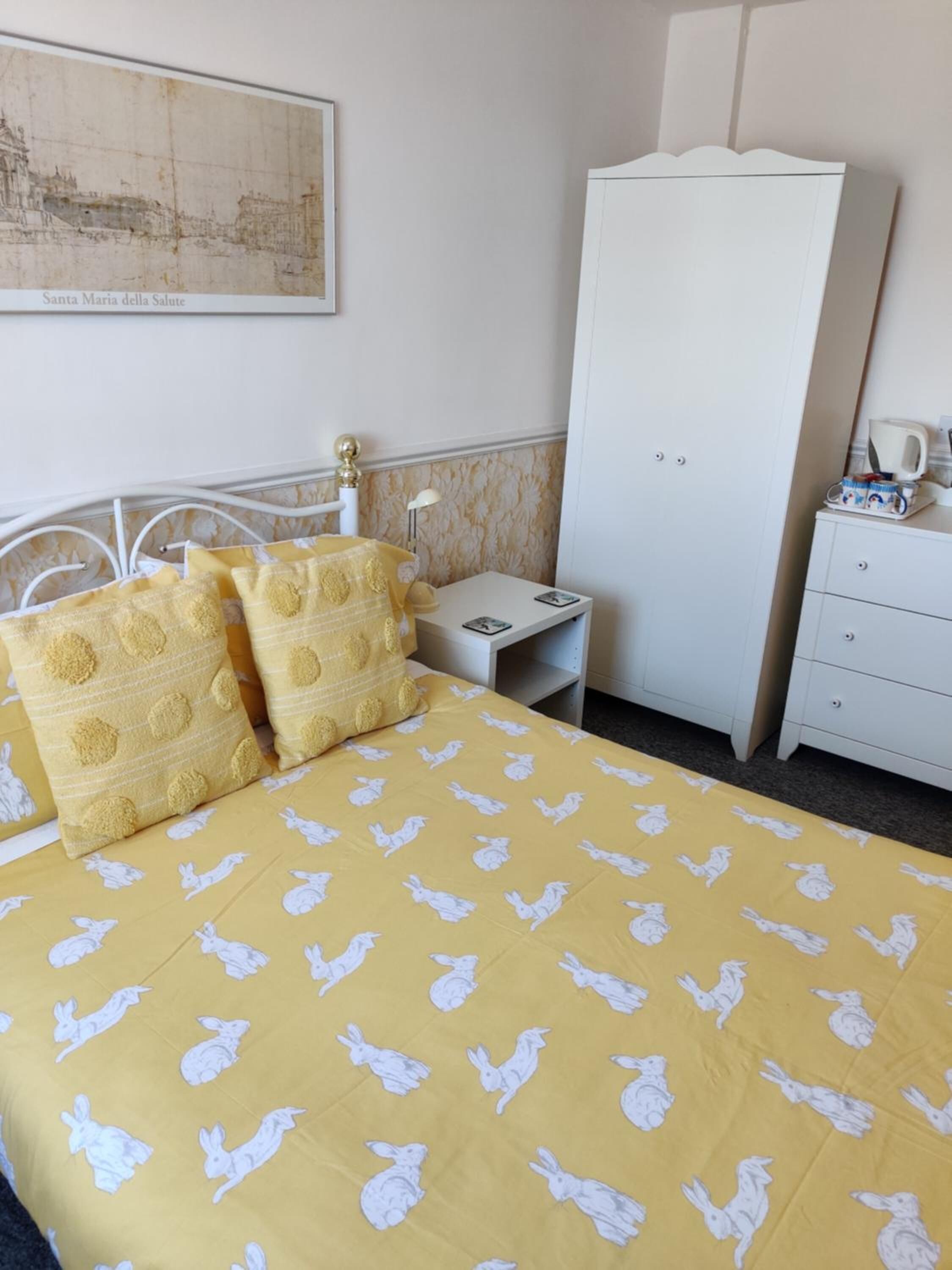 Quarto de hotel com cama coberta por um edredom amarelo com estampas de coelhos brancos, duas almofadas com design amarelo e branco, cabeceira metálica branca, guarda-roupa branco e uma pequena mesa de cabeceira com luminária e xícara.