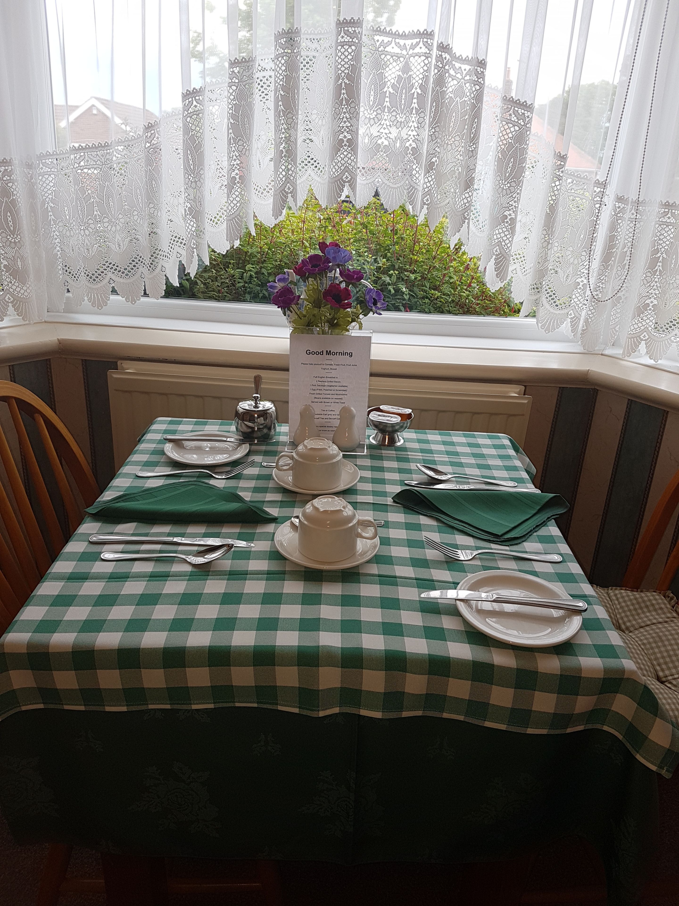A table set for breakfast with a green and white checkered tablecloth, a vase of flowers, and a menu card.
