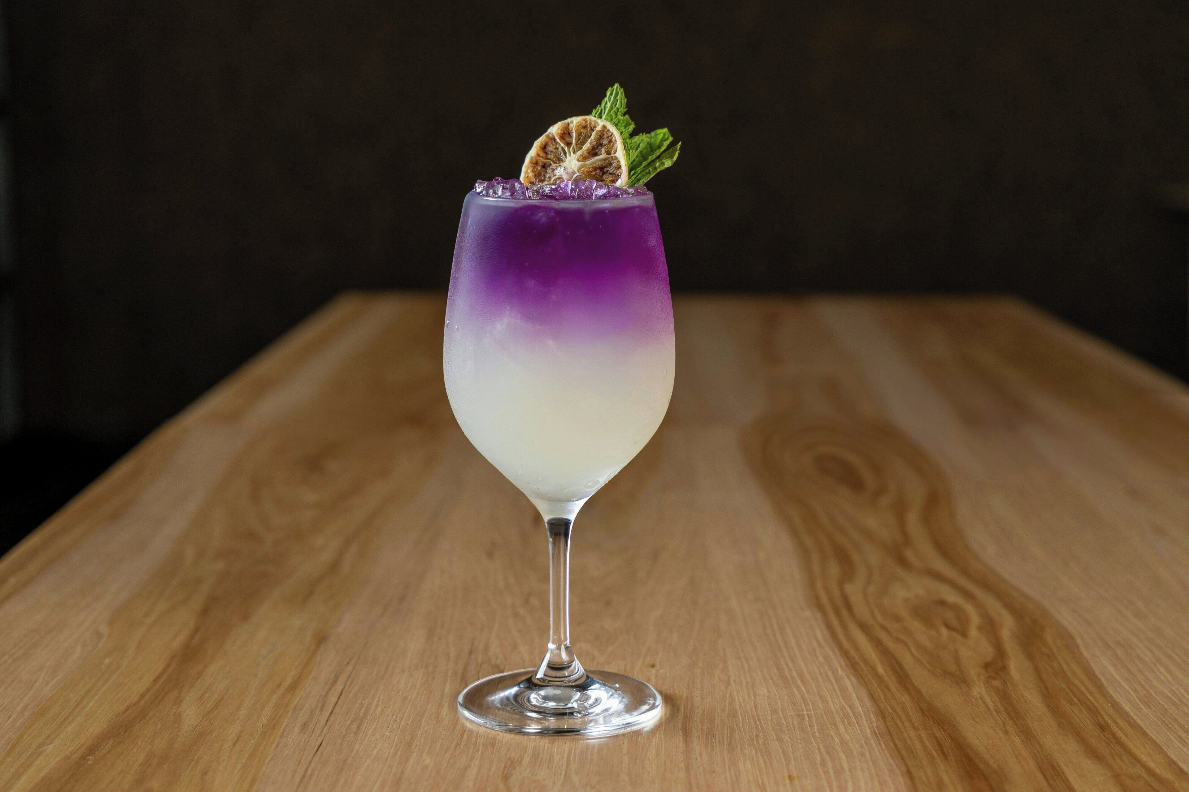 A cocktail in a stemmed glass with a gradient color effect, garnished with a slice of citrus and a mint leaf, on a wooden table.