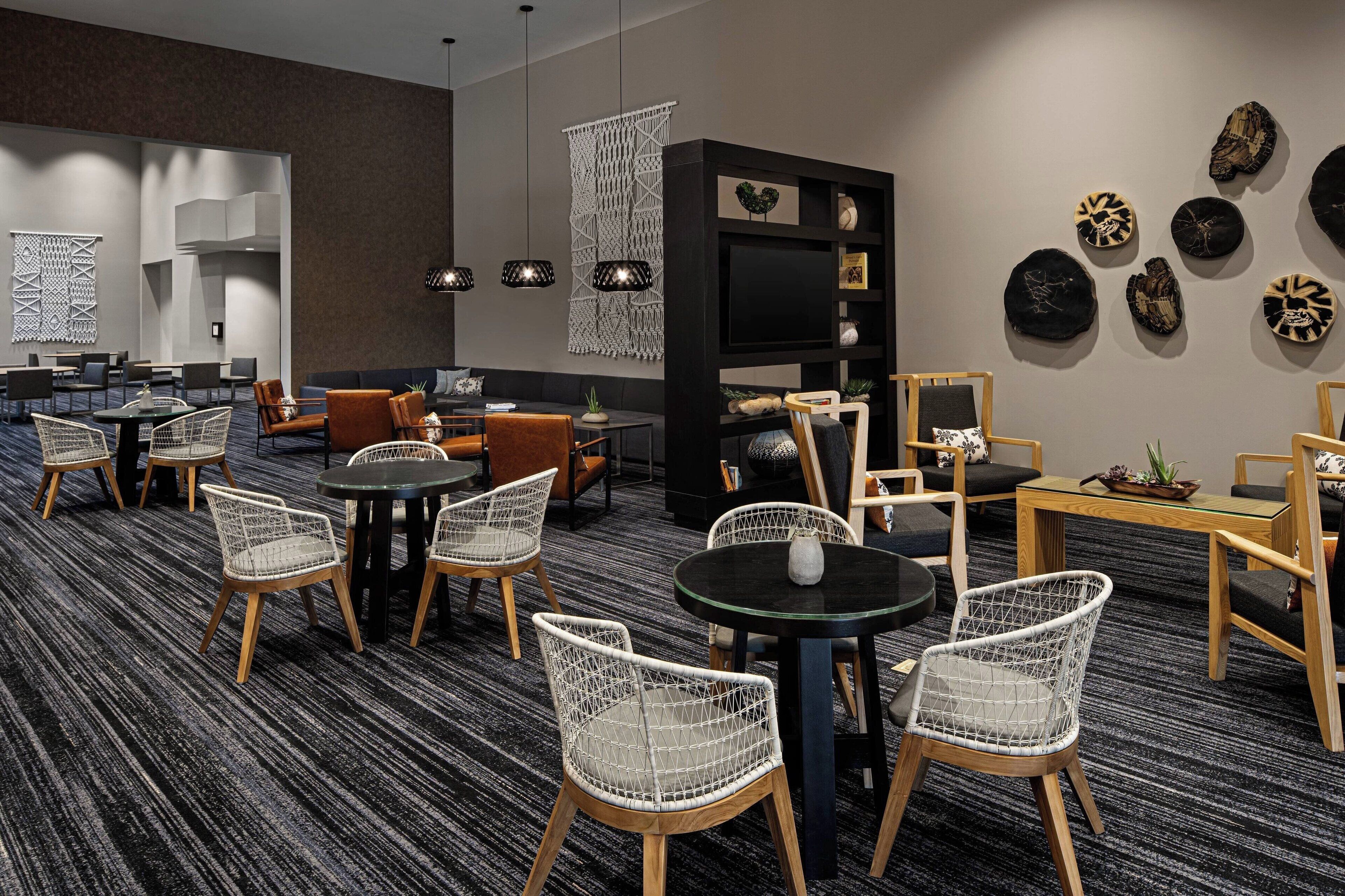 Eine moderne Hotelhalle mit verschiedenen Sitzgelegenheiten, darunter Korbstühle, Sofas und Tische. An den Wänden befinden sich dekorative Kunstwerke und ein großer Flachbildfernseher.