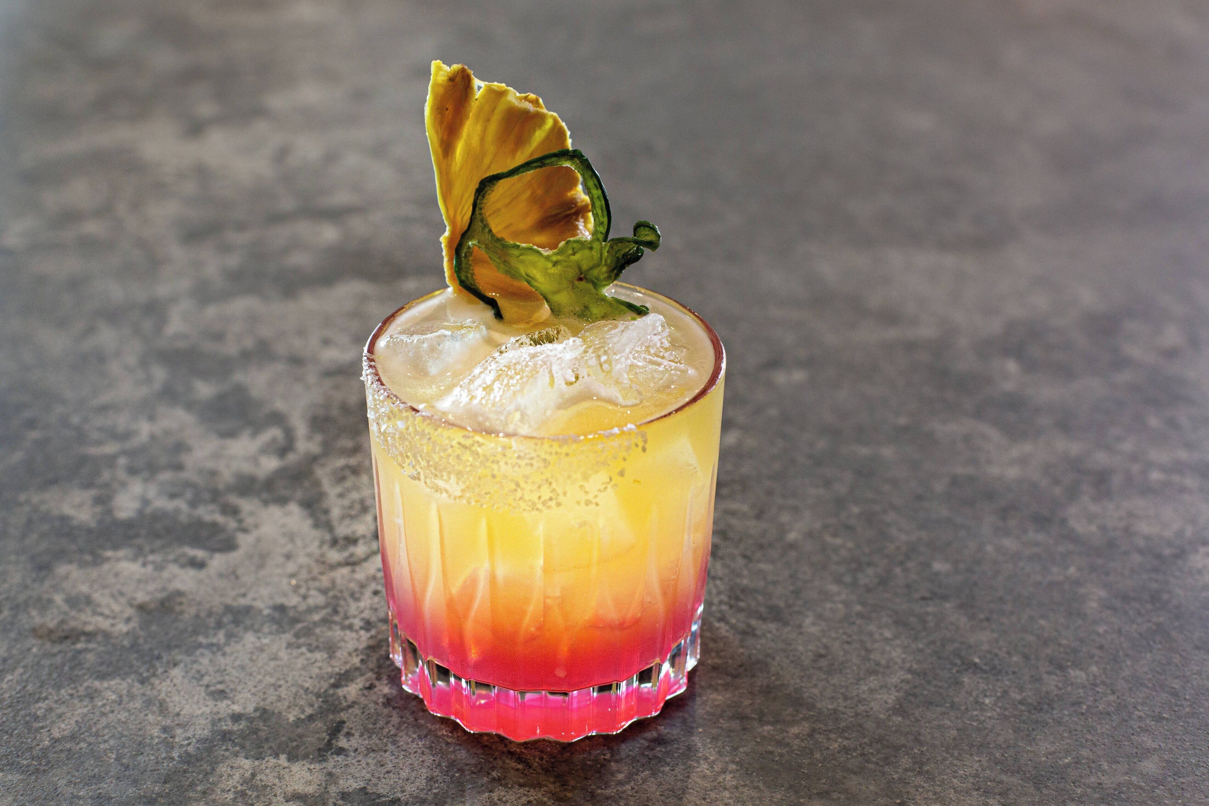 A cocktail in a glass with a gradient color from yellow to red, ice cubes, and a garnish on a textured surface.