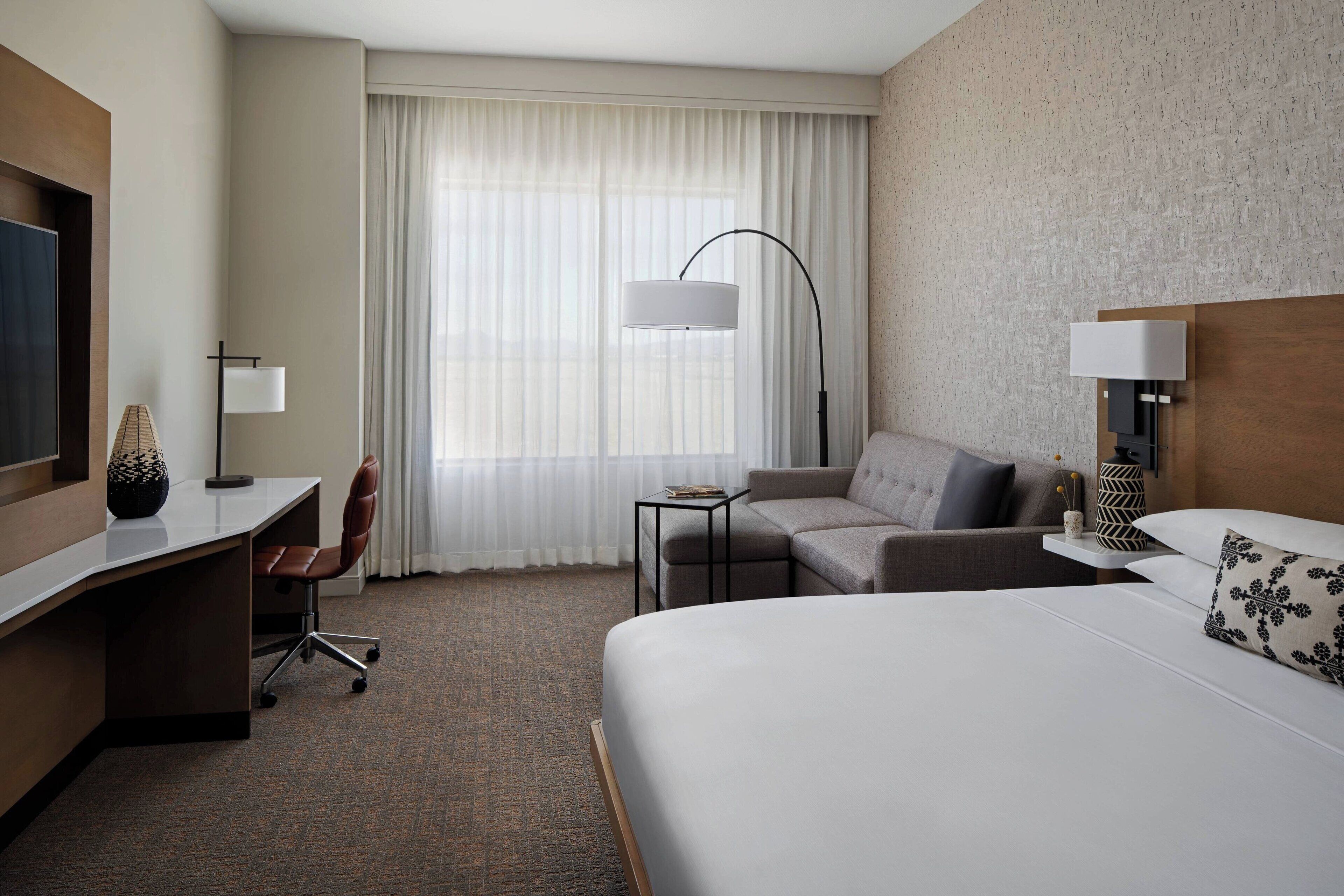 A hotel room with a large bed, a desk with a chair, a sofa, a small table, and a TV.