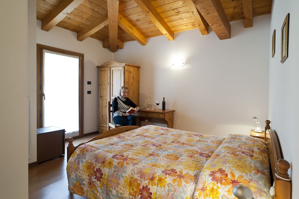 A bedroom with a wooden ceiling, a bed with a floral bedspread, a wooden desk, a cabinet, and a nightstand with a lamp.