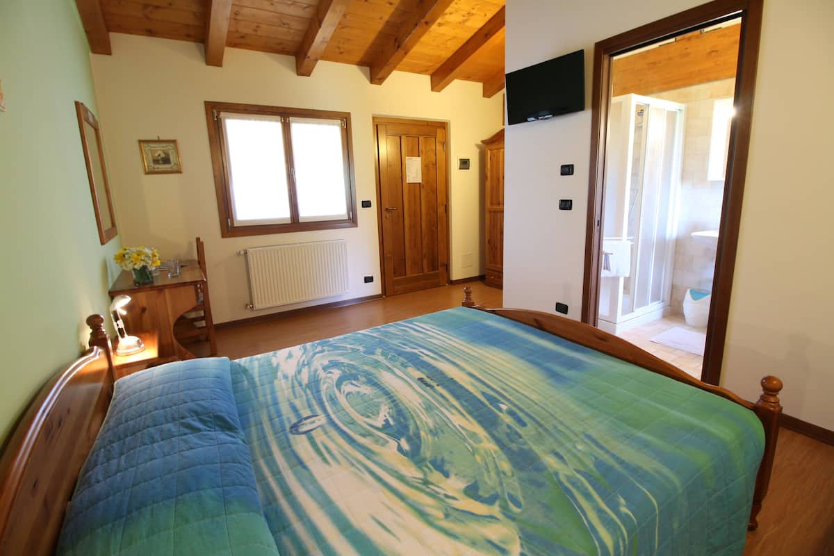 A bedroom with a large bed, wooden ceiling beams, a window, a door, and a view of a bathroom.