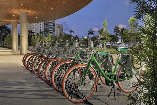 Una hilera de bicicletas verdes con aros naranja estacionadas bajo una zona cubierta con pilares de concreto.