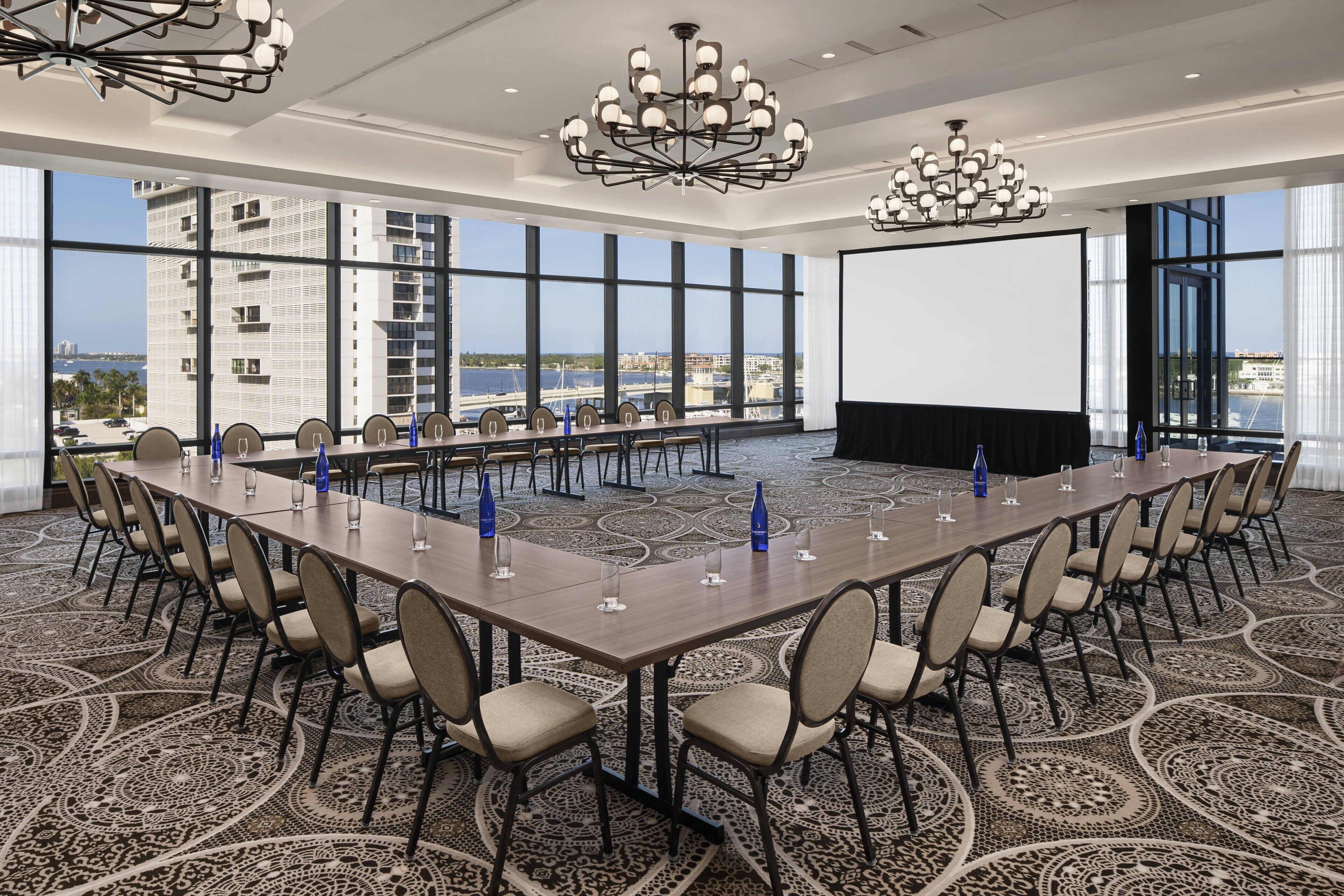 A conference room with a long table, chairs, a projector screen, and a view of a cityscape through large windows.