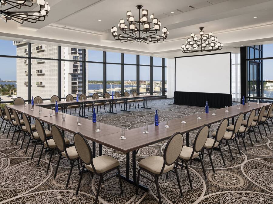 A conference room with a long table, chairs, a projector screen, and a view of a cityscape through large windows.