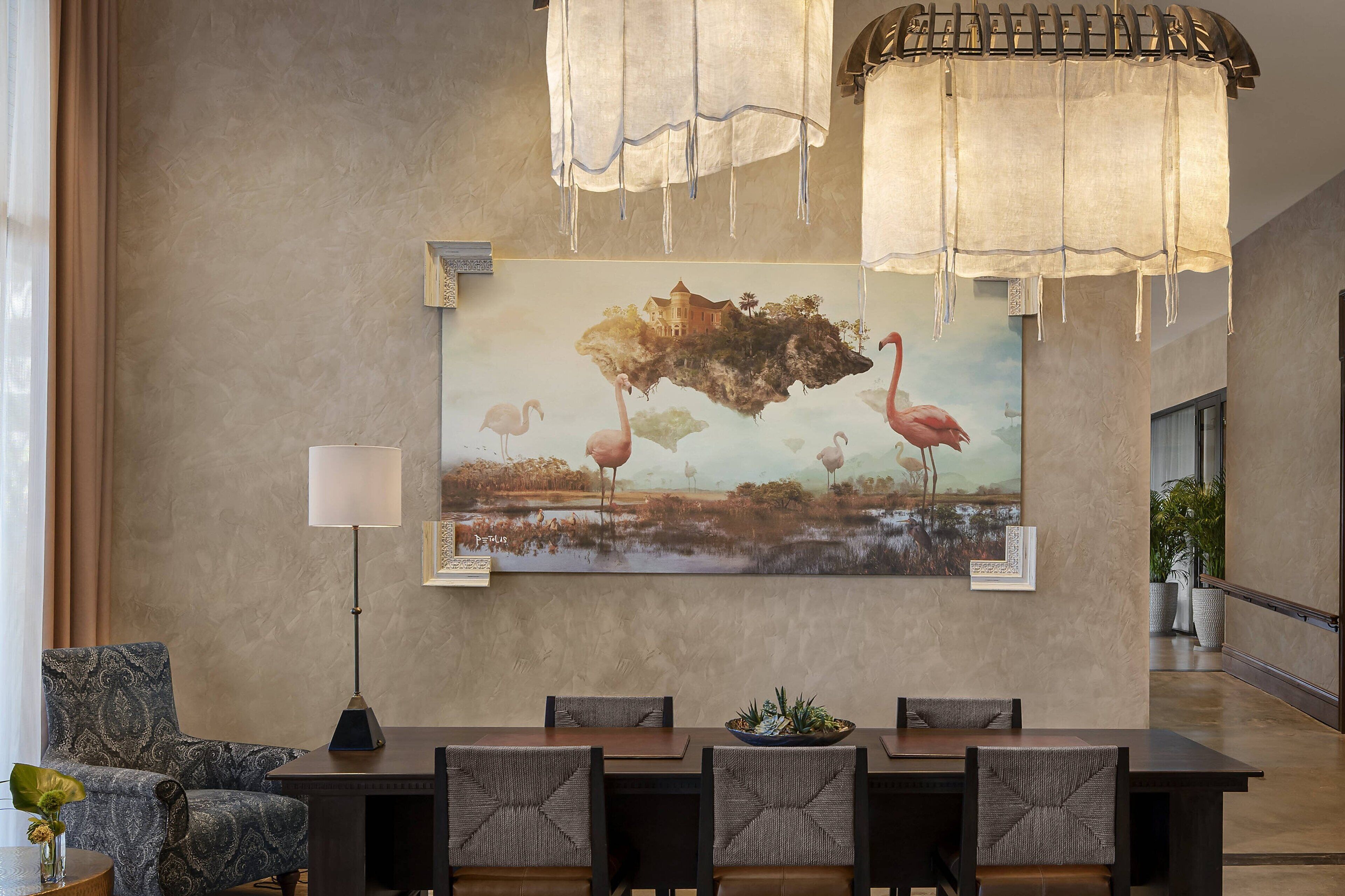 A modern dining area with a large painting of flamingos and a landscape, a chandelier, and a patterned armchair.